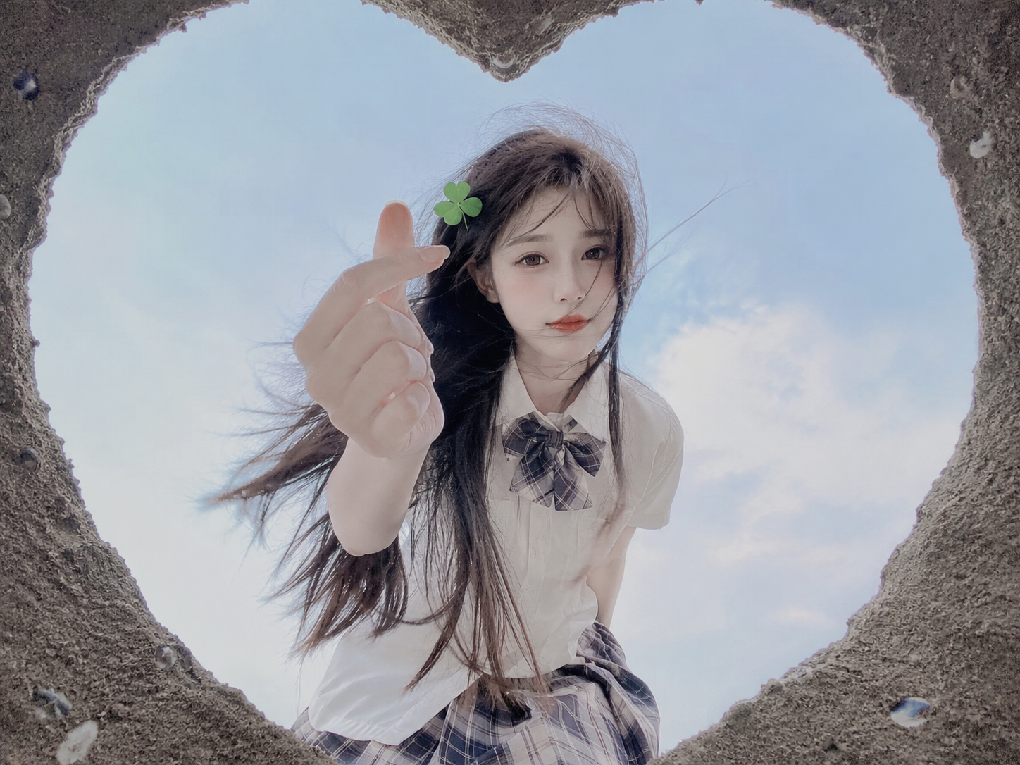 一位长发及腰、模样似洋娃娃般可爱的女生，采用超广角，体现动态美学，害羞，感受风，与镜头互动，人像摄影，日常快照风格，非精心构图或打光，小上围，妖冶少女，冷白皮，瓜子脸尖下巴，皮肤白皙，底妆通透，肤色均匀自然，纯欲风，ccd拍照感，一个沙子挖成的心形沙坑，不显示“nunu”这个词, 头发披散在肩头，右肩搭着一半头发，发间别着四叶草夹子。她穿着韩国学生装，打着漂亮的领。女人伸出右手打响指，手指白皙。从心形坑里底部的视角拍摄，画面带有氛围感，女人站在陆地上，好奇地伸手，眼神中透着一丝灵动。